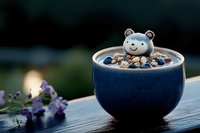 Cute bear-shaped latte art in blue ceramic bowl with nuts and berries, served on wooden table with purple flowers, moody dark background.