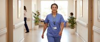Female healthcare worker in blue scrubs walking through modern hospital corridor with colleagues in background.
