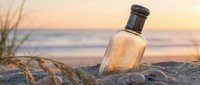 Vintage glass bottle with cork stopper resting on sandy beach at golden hour sunset. Ocean waves and coastal grass create romantic seaside atmosphere for travel concepts.