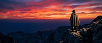 Silhouetted figure standing on rocky mountain peak at dramatic sunset with vibrant orange and red sky, suitable for inspirational and travel content.