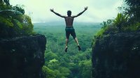 Young African American man jumping between cliffs over lush green rainforest valley, arms spread wide in freedom expression against misty mountain backdrop.