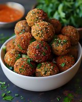 Crispy golden meatballs garnished with fresh herbs and sesame seeds served in white bowl with dipping sauce, fresh parsley scattered around on dark background.