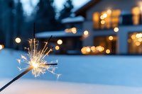 Lit sparkler against snowy winter landscape with blurred house lights and evergreen trees in background, celebrating holiday season outdoors.