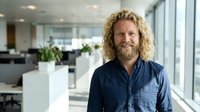 Confident middle-aged Caucasian businessman with curly blonde hair and beard smiling in modern office workspace with natural lighting and plants.