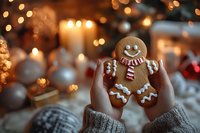 Hands holding decorated gingerbread man cookie with warm bokeh lights and candles creating cozy Christmas holiday atmosphere for seasonal baking celebrations.