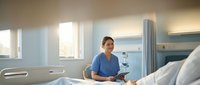 Female healthcare professional in blue scrubs reviewing patient chart in modern hospital room with natural lighting and medical equipment.