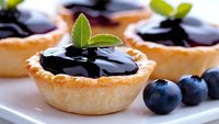 Mini tarts filled with dark blueberry jam and fresh berries, garnished with mint leaves on white plate, close up view of homemade pastry desserts.
