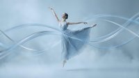 Young Caucasian female ballet dancer in flowing blue dress performing graceful leap with ethereal light trails creating magical motion effect against soft background.