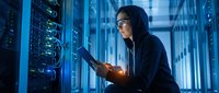 Hooded cybercriminal hacker using tablet computer in dark server room with glowing blue lights. Cybersecurity threat concept for digital crime prevention.