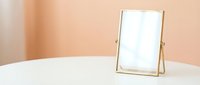 Gold metal framed mirror on white table against soft peach wall, elegant minimalist vanity decor with copy space for beauty branding.