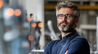 Middle-aged Caucasian businessman with gray hair and beard wearing glasses and navy sweater, standing confidently with arms crossed in modern office environment with bokeh lights.