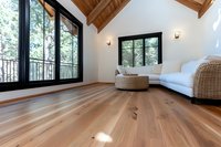 Modern attic living room with wide plank hardwood flooring, vaulted wooden ceiling, large windows, white walls, and rattan furniture creating bright airy space.