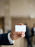 Businessman in dark suit holding blank white business card at corporate event with blurred colleagues in background for networking and professional branding purposes.