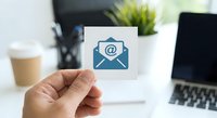 Hand holding white card with blue email envelope icon in modern office workspace with laptop and coffee cup for digital communication concepts.