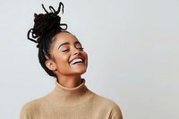Young African American woman with dreadlocks hairstyle wearing beige turtleneck sweater laughing with eyes closed against light background, expressing genuine joy and happiness.