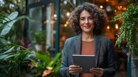 Middle-aged Mediterranean businesswoman with curly hair smiling while holding digital tablet outdoors, wearing grey blazer against blurred cafe background with plants.