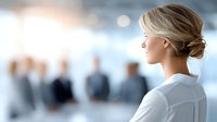 Professional blonde businesswoman in elegant updo hairstyle looking towards bright future in modern corporate office environment with blurred colleagues.