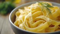 Creamy fettuccine pasta in white ceramic bowl garnished with fresh dill. Close-up view of homemade Italian noodles with rich sauce on wooden surface.
