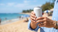 Man holding blank white beverage can at tropical beach with ocean and sandy shore in background, summer vacation and refreshment concept.