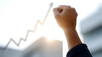 Business success concept with raised fist against bright sunlight and upward trending arrow graph symbolizing achievement, growth and victory in corporate environment.