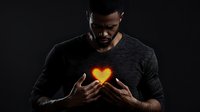 African American male holding glowing heart shape light between hands against dark background, dramatic lighting creates mystical atmosphere for wellness concepts.