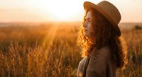 Young woman with curly hair wearing hat enjoying golden hour sunset in wheat field, peaceful countryside moment for wellness and lifestyle content.
