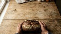 Artisan hands holding freshly baked whole grain bread loaf on rustic wooden table, showcasing homemade baking and traditional bread making craft.
