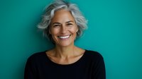 Confident mature woman with silver gray hair and genuine smile wearing black top against turquoise background, representing aging gracefully and vitality in senior years.