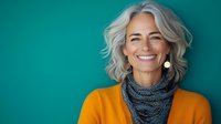 Cheerful mature woman with gray wavy hair wearing bright yellow sweater and gray scarf smiling against turquoise wall background. Natural aging and wellness concept.