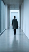 Male professional in dark business attire walking away through modern minimalist corridor with polished concrete floor and bright natural light, representing corporate ambition and forward momentum.