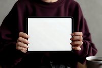 Person in burgundy sweater holding blank tablet with white screen mockup against neutral gray background, coffee cup visible on table.