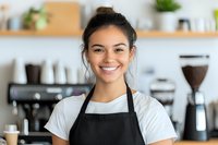 Young Asian female barista wearing black apron smiling warmly at camera in modern coffee shop interior with coffee equipment and blurred background. Professional service industry portrait.