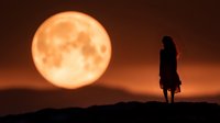 Silhouette of young woman standing on hill against dramatic orange full moon at sunset, creating mysterious atmospheric scene with long hair flowing in breeze.