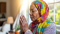 African American Muslim woman in colorful hijab praying peacefully by window, eyes closed, hands clasped in spiritual meditation pose showing religious devotion.