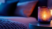 Glowing blue glass candle holder with honeycomb pattern creates cozy atmosphere on bedside table with knitted blanket in dark bedroom setting.