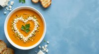 Creamy orange soup with heart-shaped herb garnish and seeds in white bowl, served with crusty bread on blue background for healthy comfort food recipes.