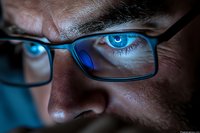Close up view of intense blue eyes behind glasses reflecting computer screen light, dramatic dark lighting creates moody atmosphere for digital lifestyle concept.