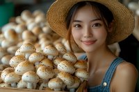 Young Asian woman in straw hat and denim overalls holding fresh baked buns at bakery, warm natural lighting creates inviting atmosphere for food photography.