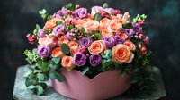 Fresh floral arrangement with peach and purple roses in pink ceramic bowl, mixed with delicate greenery against dark background, romantic gift composition.