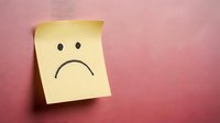 Yellow sticky note with sad face emoticon on pink textured background expressing negative emotions and disappointment for mental health concepts.