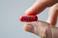 Red capsule pill with power text held between fingers against light background, representing strength, enhancement and motivation concept.