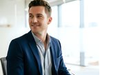 Caucasian male professional in navy blazer and light blue shirt smiling at camera in modern office with bright windows, suitable for corporate, business, and leadership concepts.