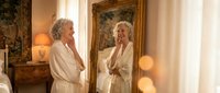 Elegant senior woman in luxurious bathrobe admiring reflection in ornate golden mirror during morning skincare routine in upscale bedroom interior.