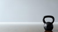 Black kettlebell on wooden floor against white wall in minimalist gym setting for fitness and strength training workouts.