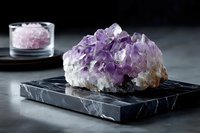 Raw amethyst crystal cluster on black marble base with pink salt in glass container against dark background for wellness and home decor.