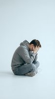 Young man in gray hoodie sitting on floor holding head in hands showing depression, stress, anxiety, mental health struggles on white background.