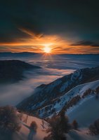 Dramatic winter sunset over snow-covered mountain peaks with low-lying clouds creating mystical atmosphere in valley below, orange sky and sunburst above foggy landscape.
