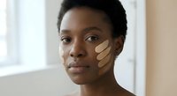 Young African American woman applying foundation makeup with finger strokes on cheek in natural light for skincare and beauty routine.