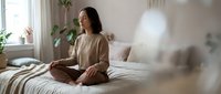 Young Asian woman practicing meditation in lotus position on bed in bright modern bedroom with plants creating peaceful morning wellness routine.
