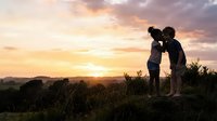Mother and young child embracing outdoors at sunset, silhouetted against golden sky with rolling hills and vegetation, capturing tender family bonding moment during golden hour.